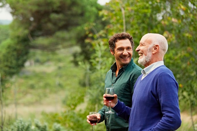 Two men smiling and talking while holding glasses of red wine outdoors among green trees.