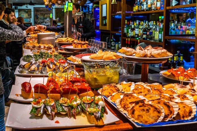 Colorful pintxos displayed on platters along a bustling bar counter in San Sebastián.