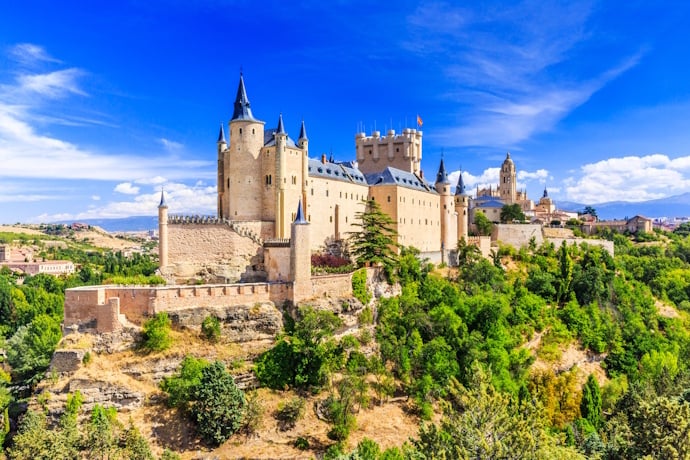 Panoramic view of Segovia’s Alcázar castle with turrets rising above green hills under a blue sky.