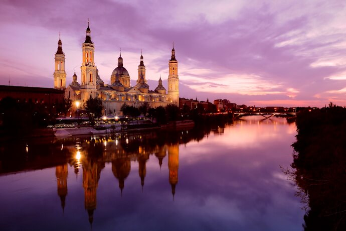 Basilica of Our Lady of the Pillar in Zaragoza illuminated at dusk, reflected in the calm Ebro River under a purple sky.