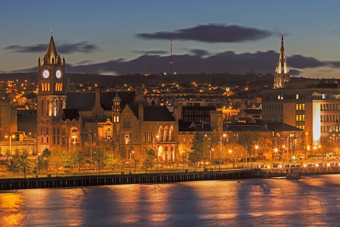 Illuminated Guildhall and city skyline along the River Foyle at dusk in Derry, Northern Ireland.