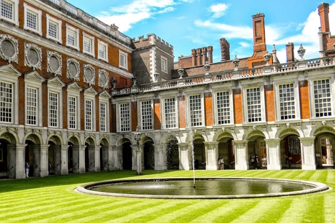 Hampton Court Palace, with historic brick walls and towers in the background and a fountain in the center.