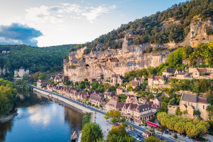 Riverside village in the Dordogne Valley beneath towering limestone cliffs surrounded by forests.