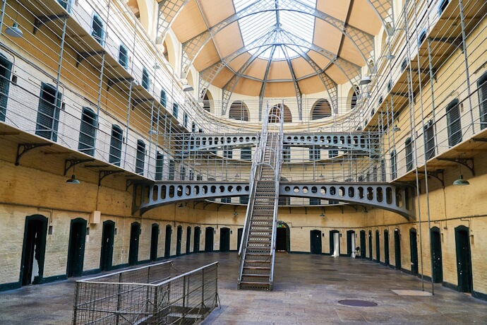 The interior of the historic Kilmainham Gaol prison featuring multiple tiers of cells, iron staircases, and walkways surrounding a large central atrium topped with a glass dome ceiling.