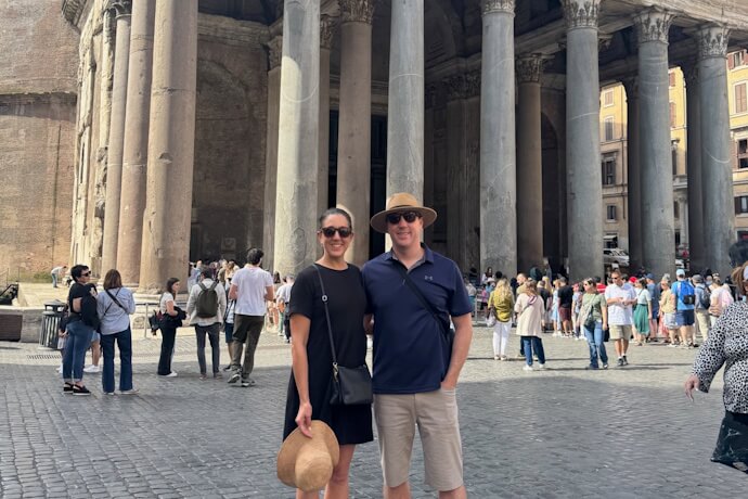 Couple standing in front of the Pantheon in Rome, Italy, posing for a photo in the piazza, where people are admiring the Roman temple