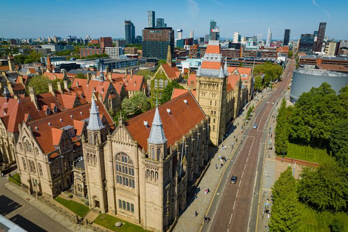 Aerial view of the University of Manchester with its red-roofed Gothic buildings along Oxford Road, framed by the modern skyline of Manchester, England