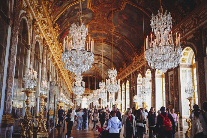 People exploring the Hall of Mirrors, a grand gallery with chandeliers and gilded decor inside the Palace of Versailles, France