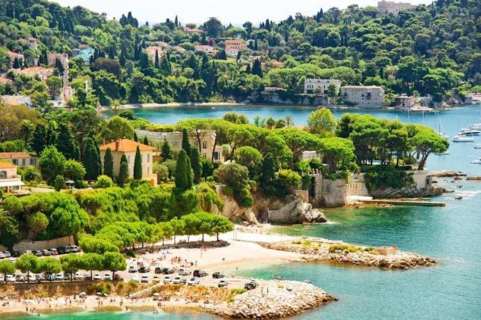 Scenic Mediterranean coastline of Villefranche-sur-Mer, France, with turquoise water, pine trees, and elegant villas near a small beach.