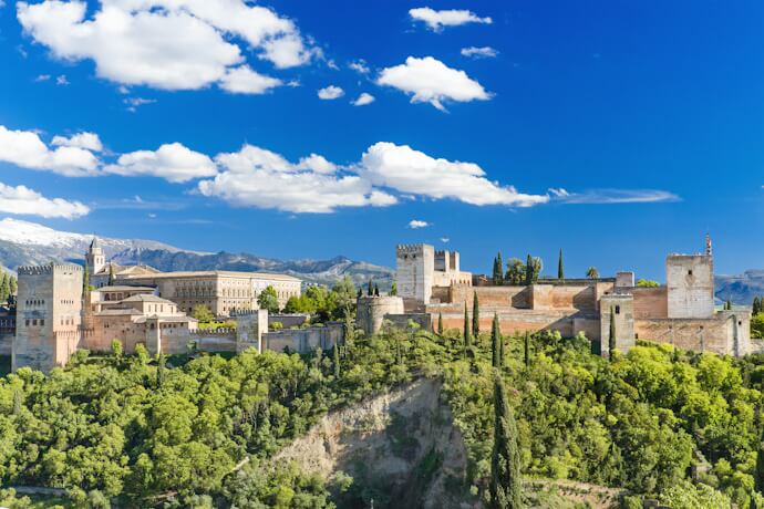 Wide view of the Alhambra palace complex in Granada, with its towers and walls rising above lush greenery and the city below