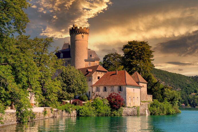 Medieval lakeside castle with tower and red-roofed buildings along the Doubs River in eastern France.