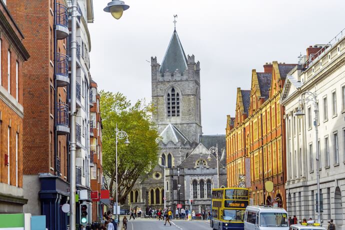 View along a busy Dublin street lined with historic buildings and a double-decker bus, leading to Christ Church Cathedral with its stone tower and spire in the background.
