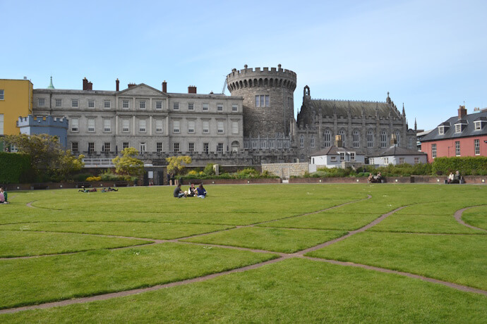 Wide lawn with people sitting on the grass in front of Dublin Castle, featuring its medieval round tower and long grey Georgian-style building