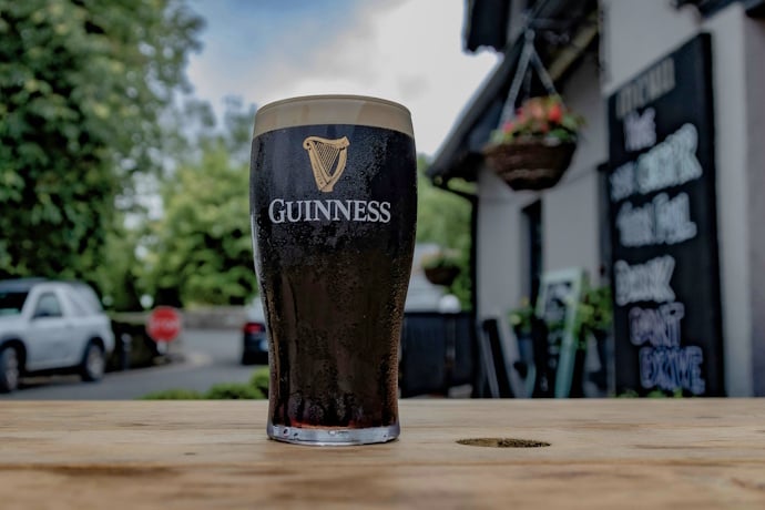 A pint of Guinness stout with its creamy head served in a branded glass, placed on a wooden outdoor table with a blurred background.
