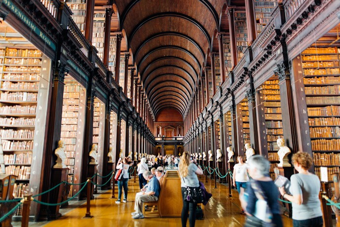The Long Room of the Old Library at Trinity College Dublin, with tall wooden shelves filled with books, arched ceiling, marble busts, and visitors exploring the historic space.