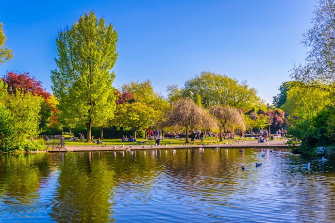 View of St. Stephen’s Green park in Dublin, with a large pond reflecting lush trees, people strolling along paths, and birds on the water