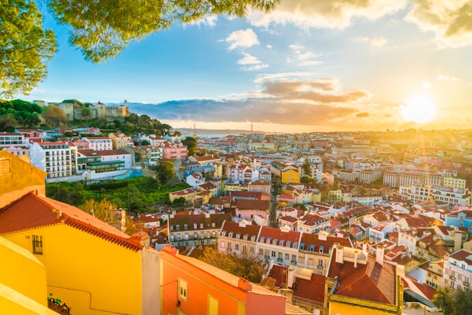 A colorful cityscape spreads across the hills, with red rooftops, pastel houses, and São Jorge Castle catching the golden light of sunset.