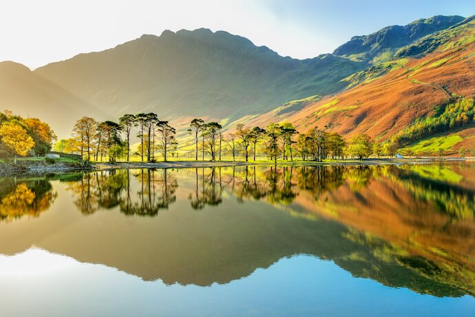 Peaceful reflection of trees and golden hills on a calm lake in the Lake District, bathed in soft sunlight.