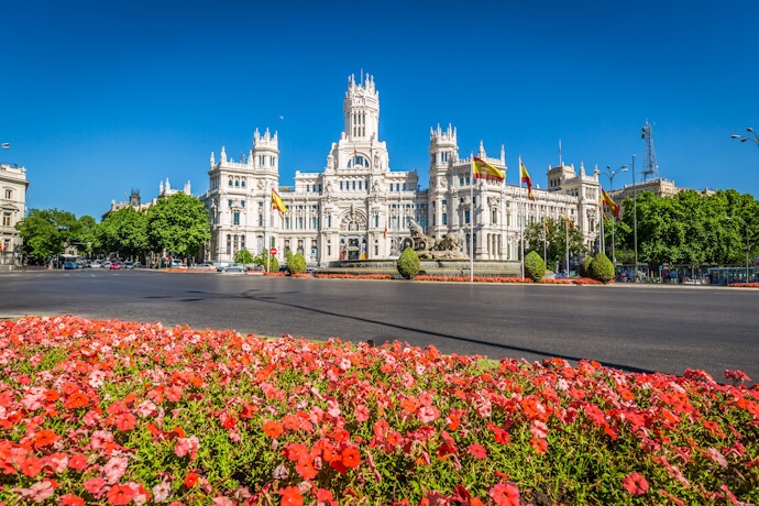 In Madrid, Spain, the white Palacio de Cibeles overlooks a broad roundabout, framed by Spanish flags, green trees, and a wide bed of red and pink flowers.