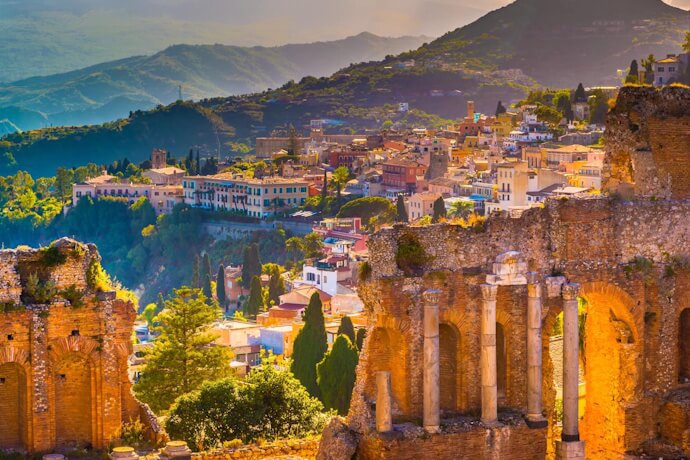 In Taormina, Sicily, ancient stone arches of the Greek Theatre frame the hillside town and layered mountains glowing in warm afternoon light.