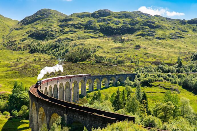 In the Scottish Highlands, a steam train crosses the curved Glenfinnan Viaduct, trailing white smoke above green valleys.