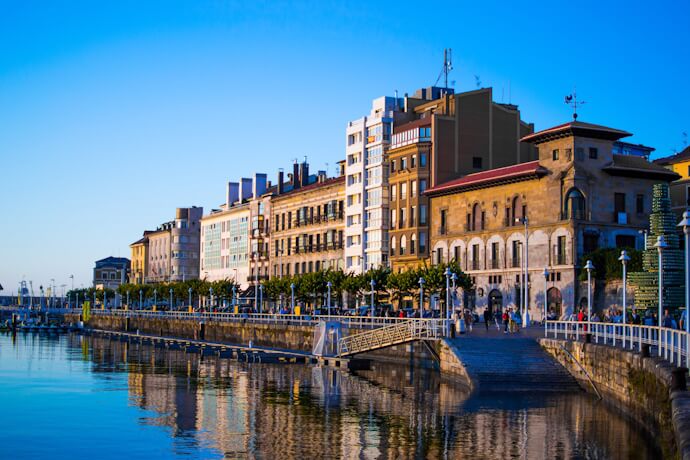 Waterfront promenade in Gijón, Spain, with historic buildings reflected in the calm harbor waters