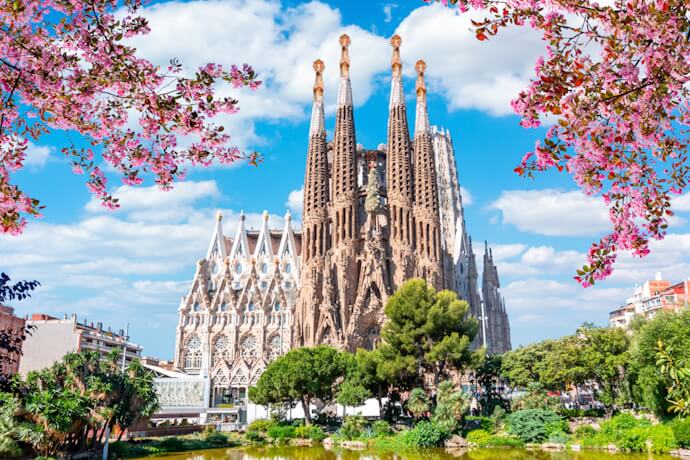 View of the Sagrada Família framed by pink blossoms, with the pond and gardens in the foreground.