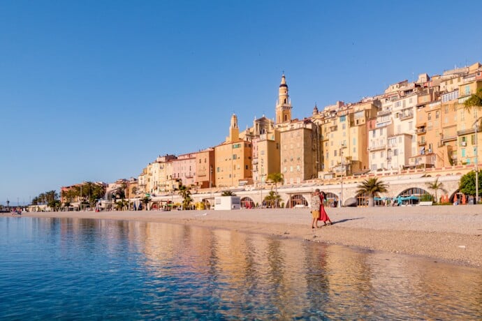 In Menton, France, pastel houses line the waterfront above a quiet beach, reflected in calm Mediterranean water.